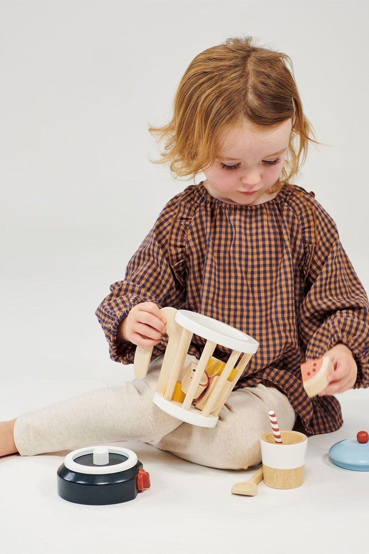 A child plays with a wooden smoothie maker toy set, featuring a blender and wooden fruit pieces.