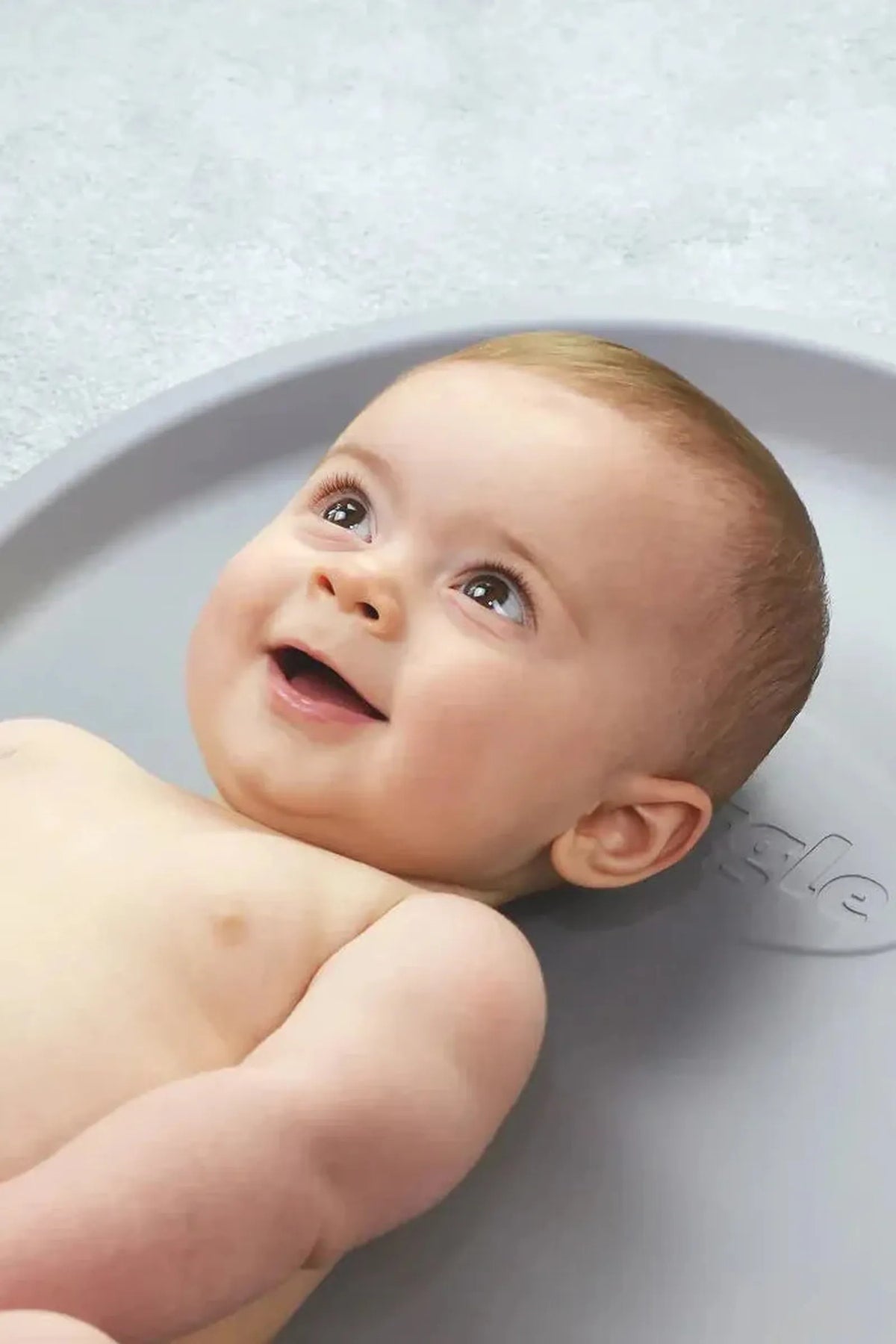 Smiling baby lying on a gray mat, looking upwards with bright eyes. The mat features subtle embossed branding.