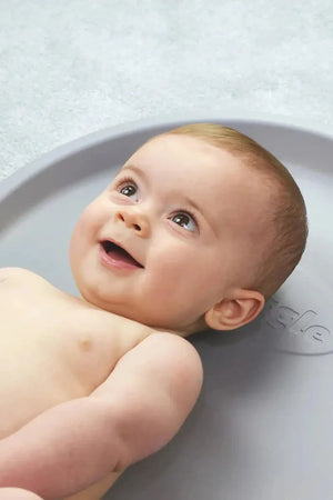 Smiling baby lying on a gray mat, looking upwards with bright eyes. The mat features subtle embossed branding.