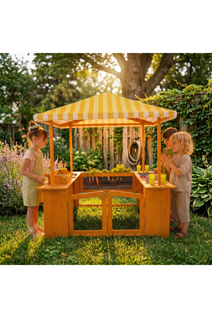 Two children play at a vibrant wooden market stand with a yellow striped canopy in a lush garden, surrounded by flowers and greenery, enjoying imaginative outdoor play.