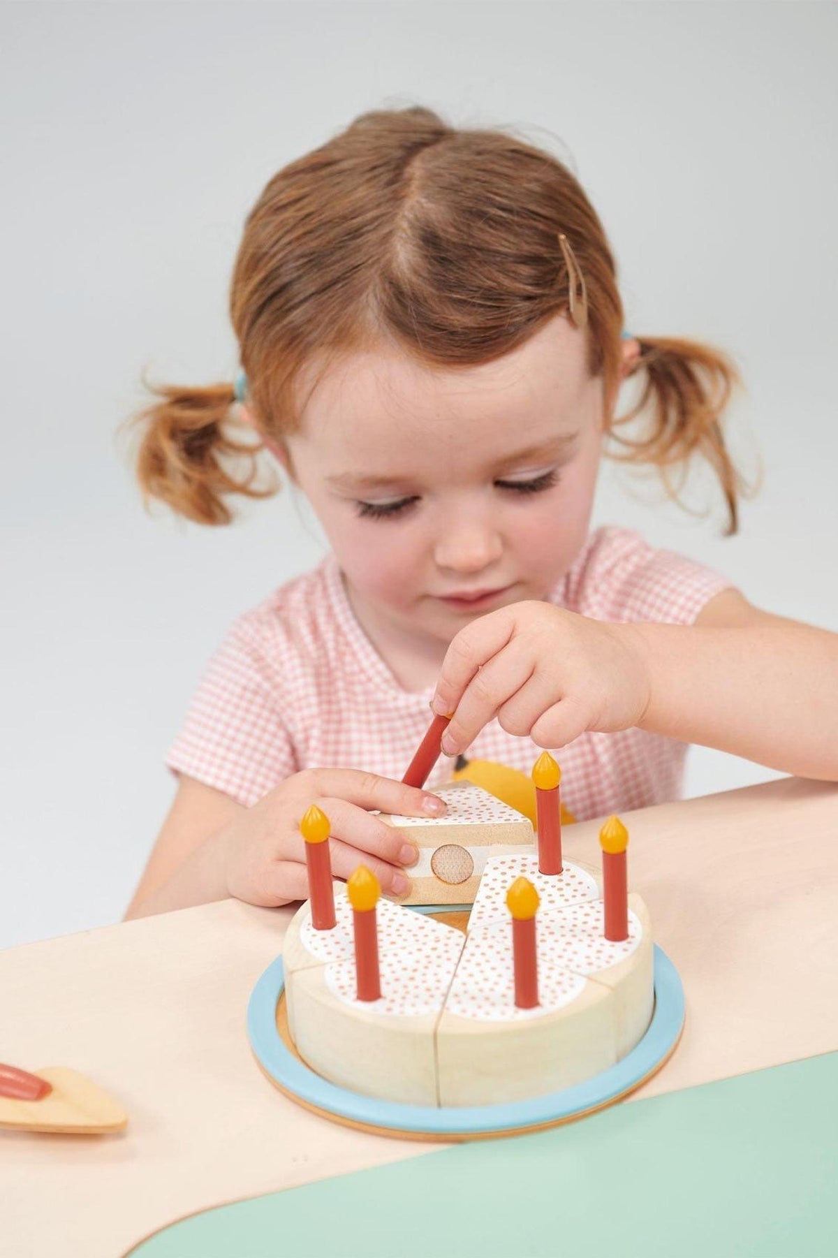 A child with pigtails plays with a wooden toy cake featuring red candles and a blue base.