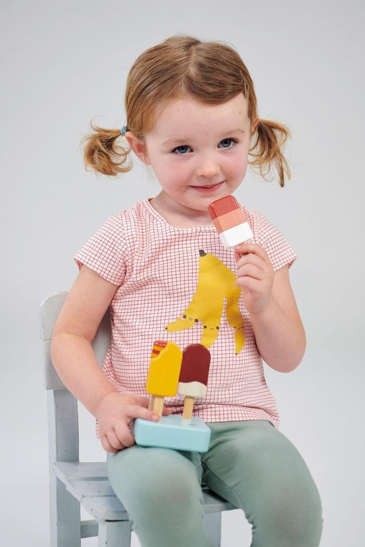 A child in a red checked shirt with a banana design holds wooden ice cream toys, seated on a chair.