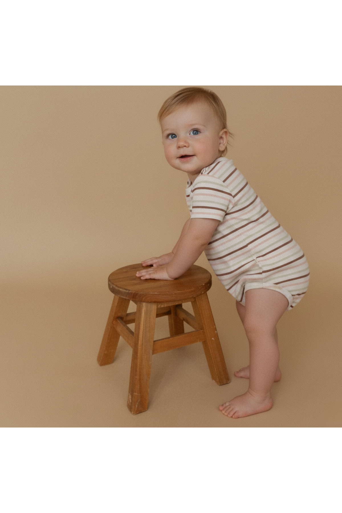A smiling baby stands beside a wooden stool, wearing a striped organic cotton romper in earthy tones. The setting features a soft, neutral background, emphasizing the rompers natural charm.