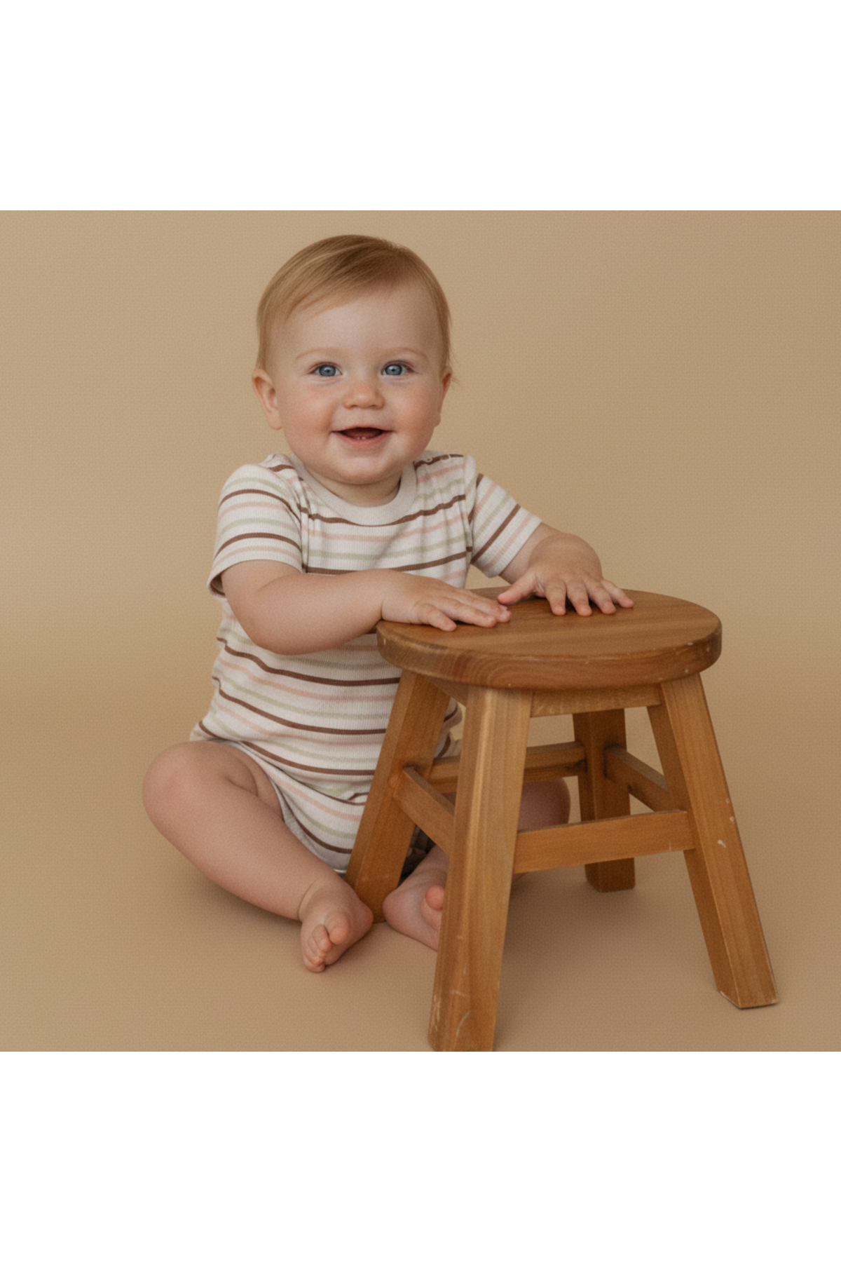 A smiling baby sits on the floor beside a wooden stool, wearing a striped short-sleeve romper in earthy tones.