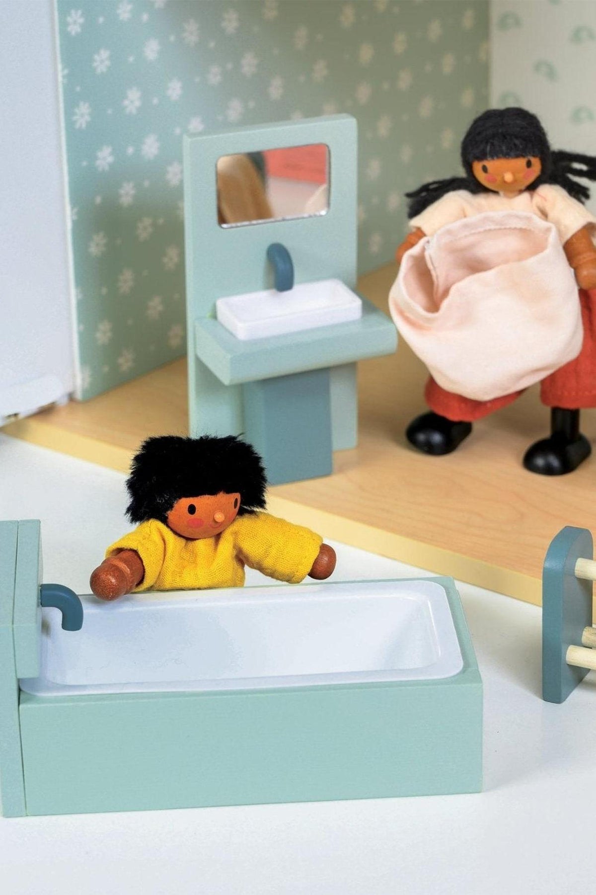 Two wooden dolls in a miniature bathroom setting with a pastel-colored bathtub and sink.