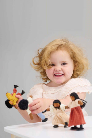 A smiling child plays with wooden dolls, featuring bendy arms and colorful knitted clothing, on a white table.