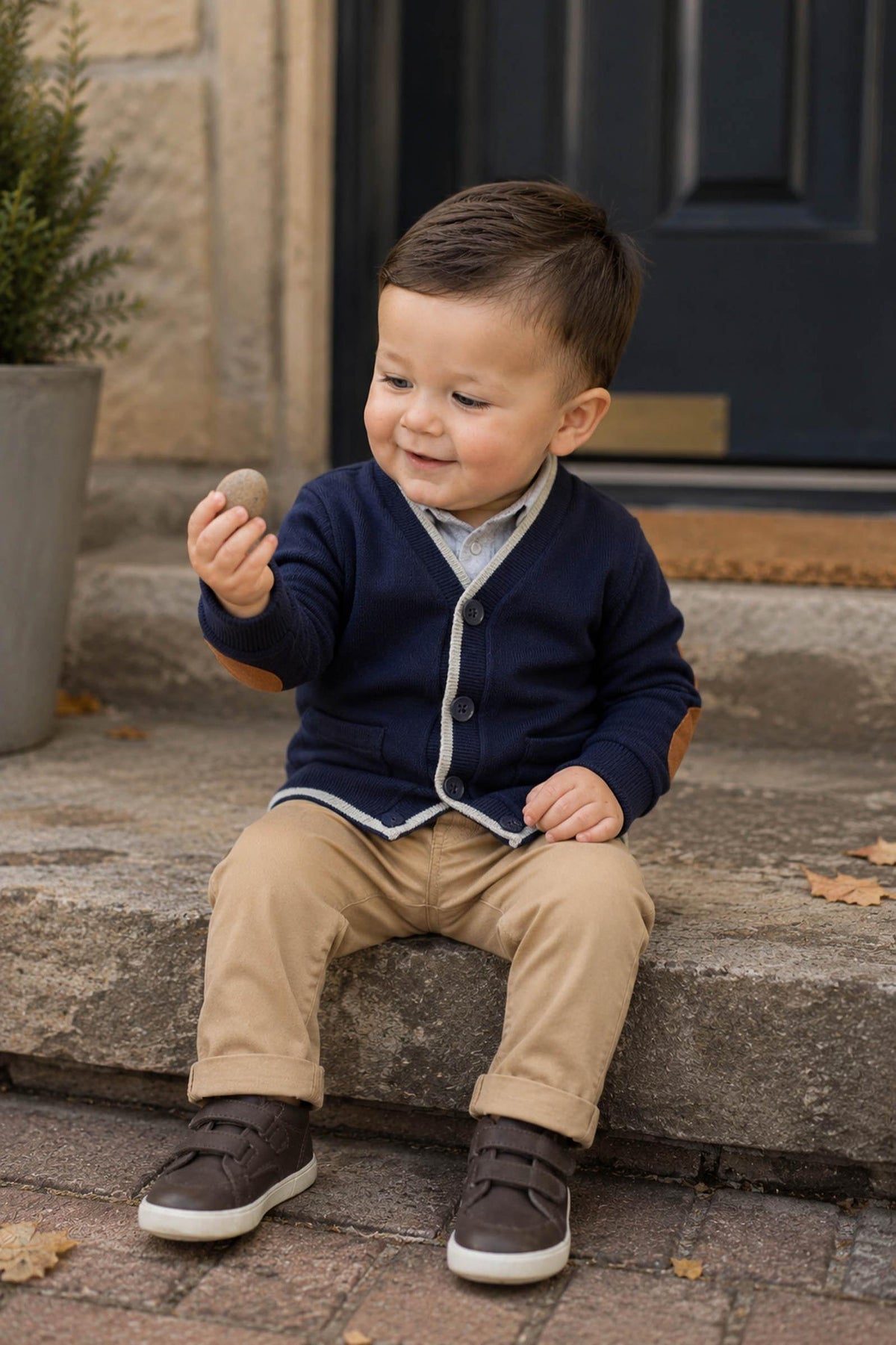 A smiling toddler sits on stone steps, wearing a navy cardigan with elbow patches, beige pants, and brown shoes. He holds a small stone, surrounded by autumn leaves and greenery.