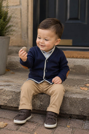 A smiling toddler sits on stone steps, wearing a navy cardigan with elbow patches, beige pants, and brown shoes. He holds a small stone, surrounded by autumn leaves and greenery.