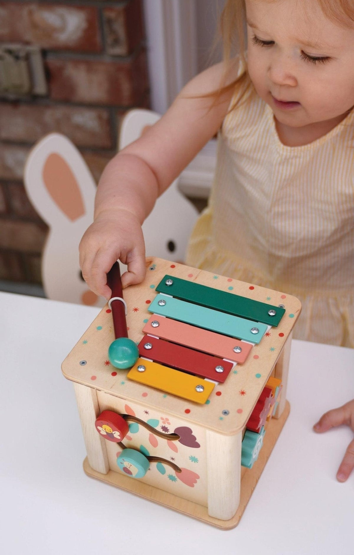 A toddler plays with a colorful xylophone on a wooden activity cube featuring gears and painted designs.