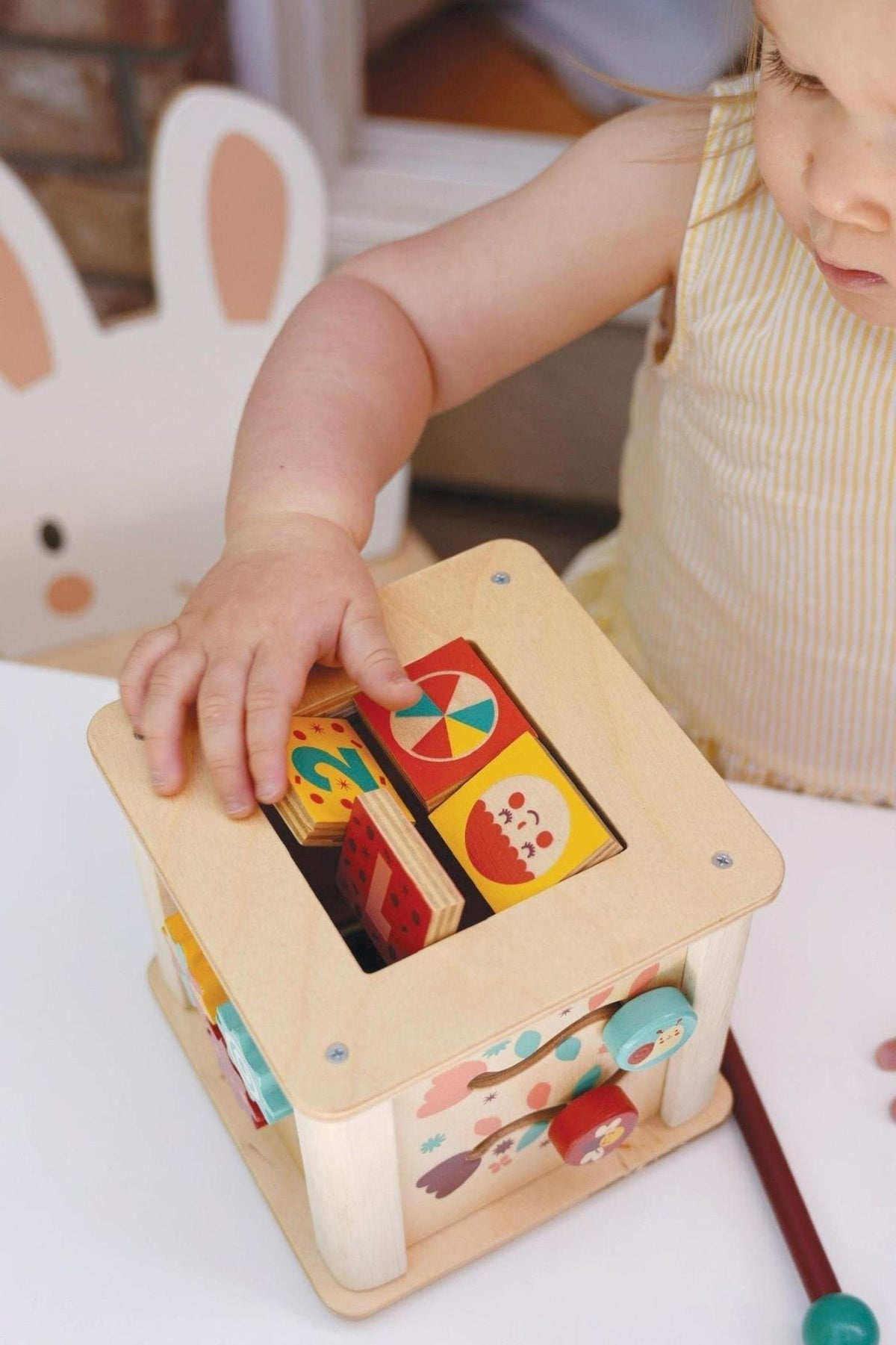 Toddler playing with a wooden activity cube featuring colorful blocks and illustrated sides on a white table.