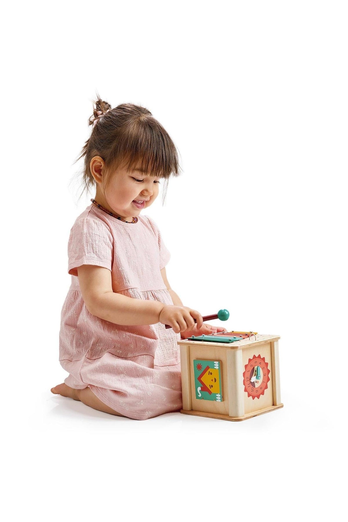 A toddler in a pink dress plays with a wooden activity cube featuring colorful designs and a xylophone.
