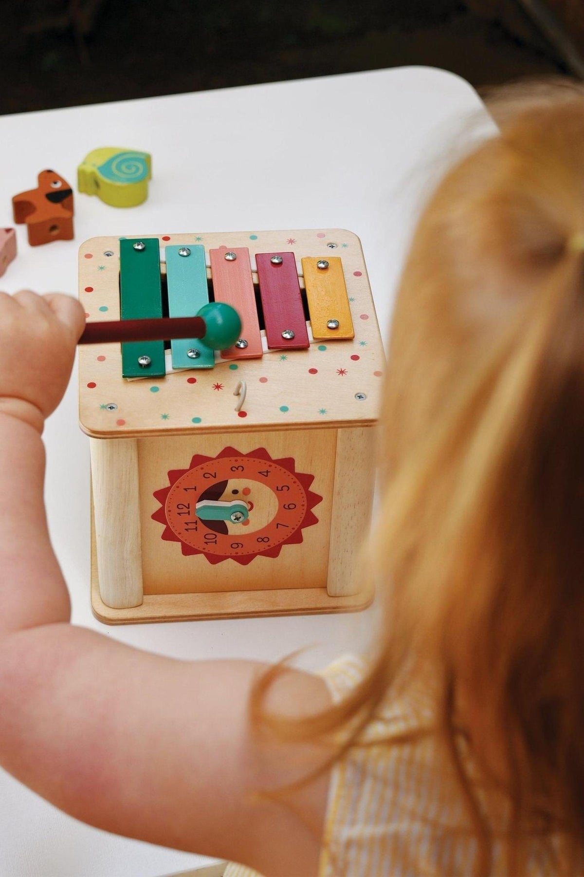 A child plays with a colorful xylophone on a wooden activity cube featuring a clock illustration and shapes.