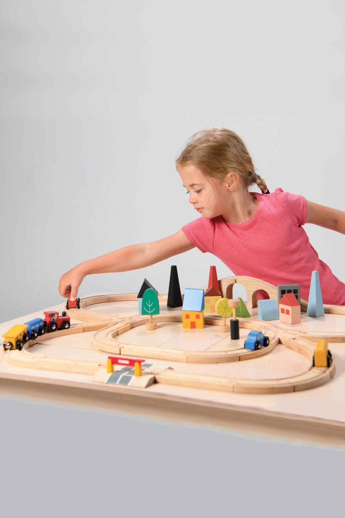 A child in a pink shirt plays with a colorful wooden train set featuring buildings and trees on a table.