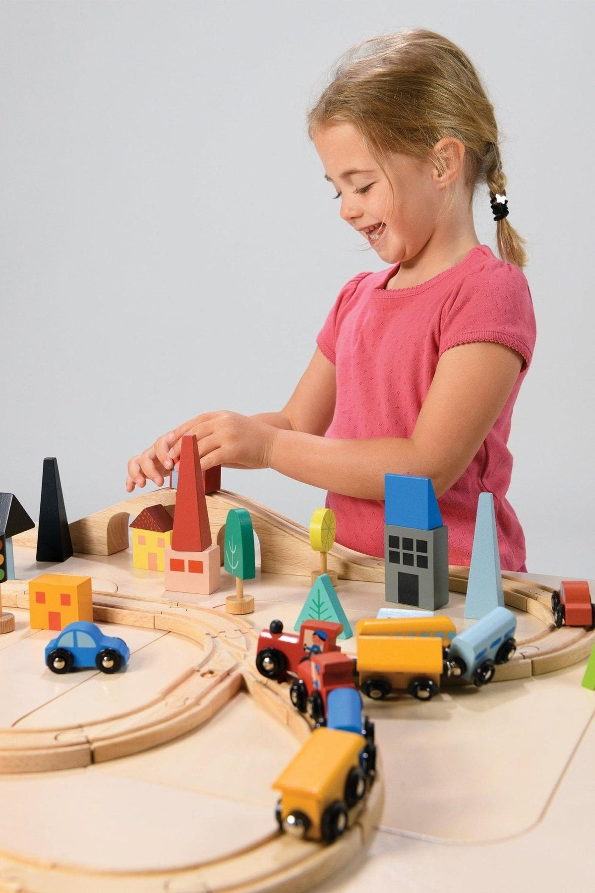 A child in a pink shirt plays with a colorful wooden train set, featuring houses and trees on a track.