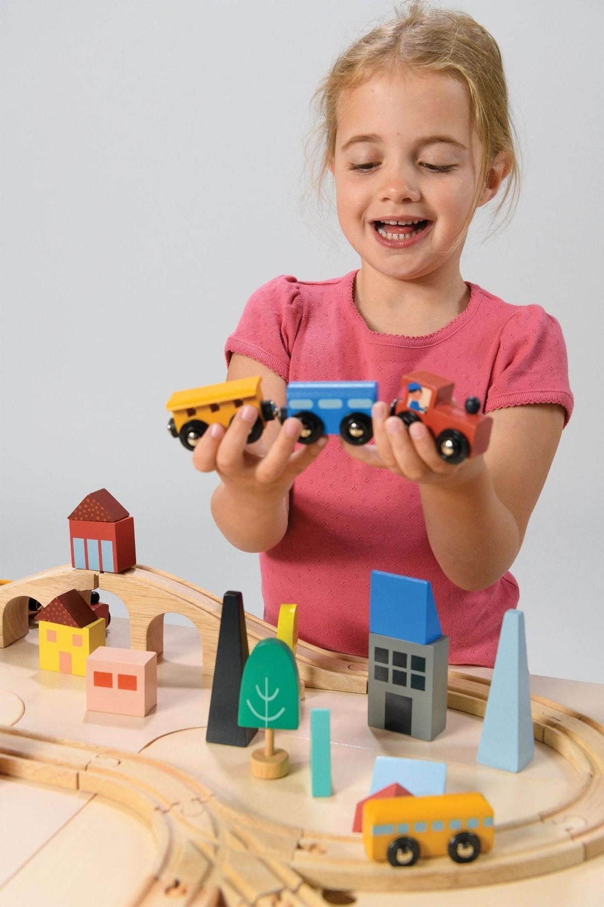 A child in a pink shirt plays with a colorful wooden train set on a wooden track with buildings.