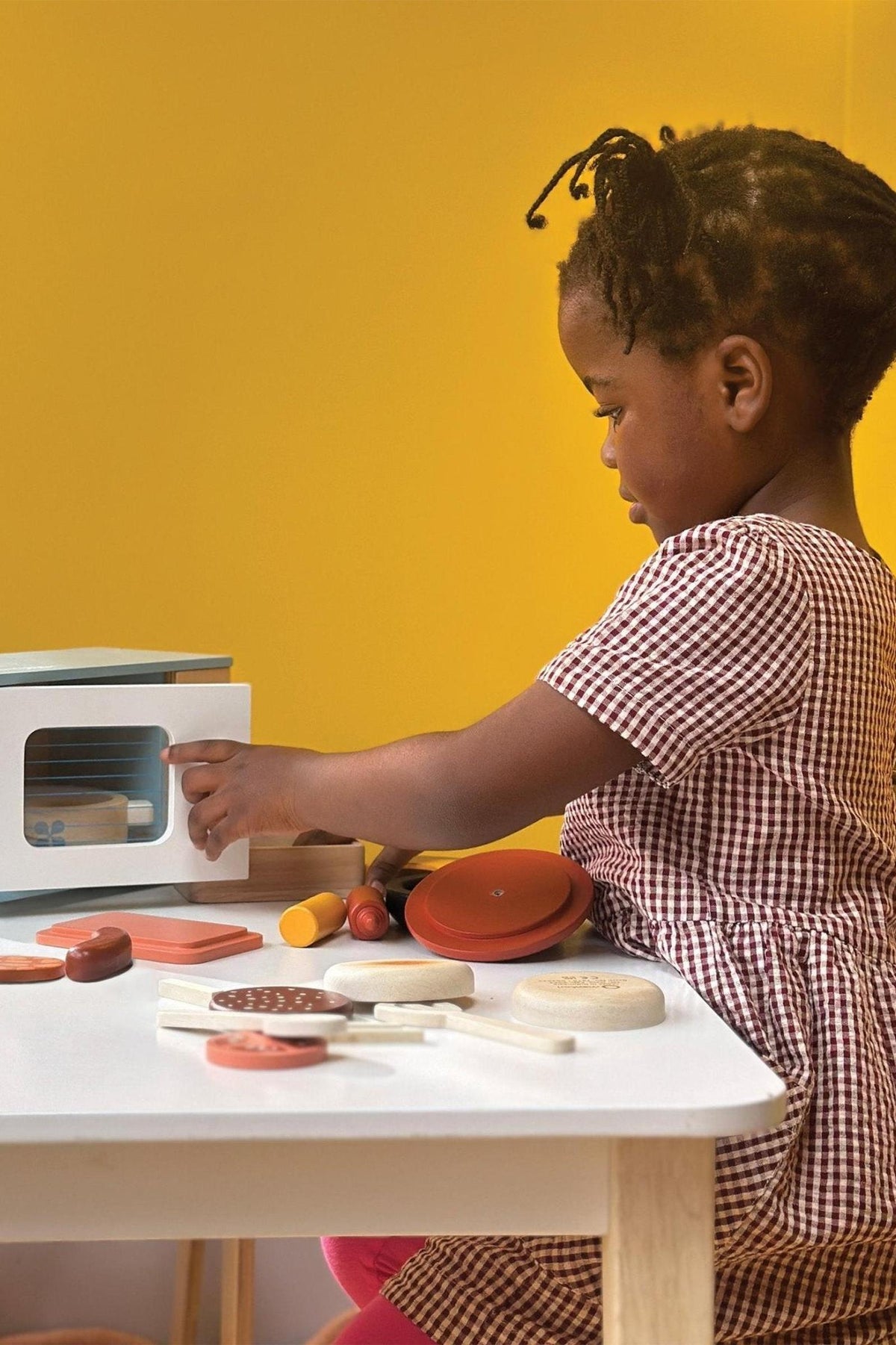 A child interacts with a white wooden toy microwave on a table with various colorful toy foods.