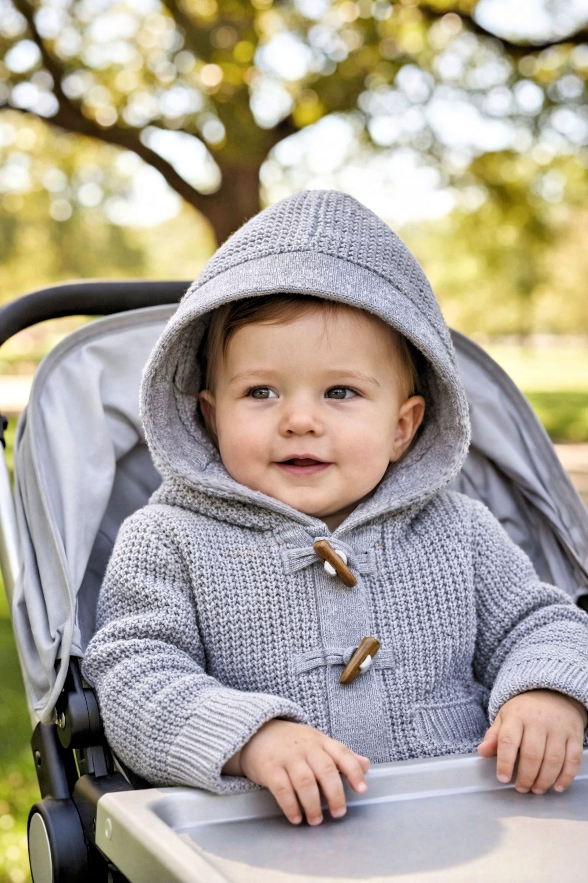 A smiling baby wears a gray hooded sweater with wooden toggles, seated in a stroller against a blurred outdoor background.