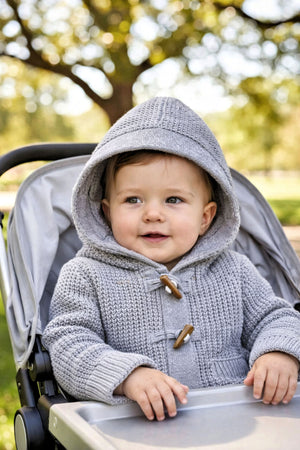 A smiling baby wears a gray hooded sweater with wooden toggles, seated in a stroller against a blurred outdoor background.