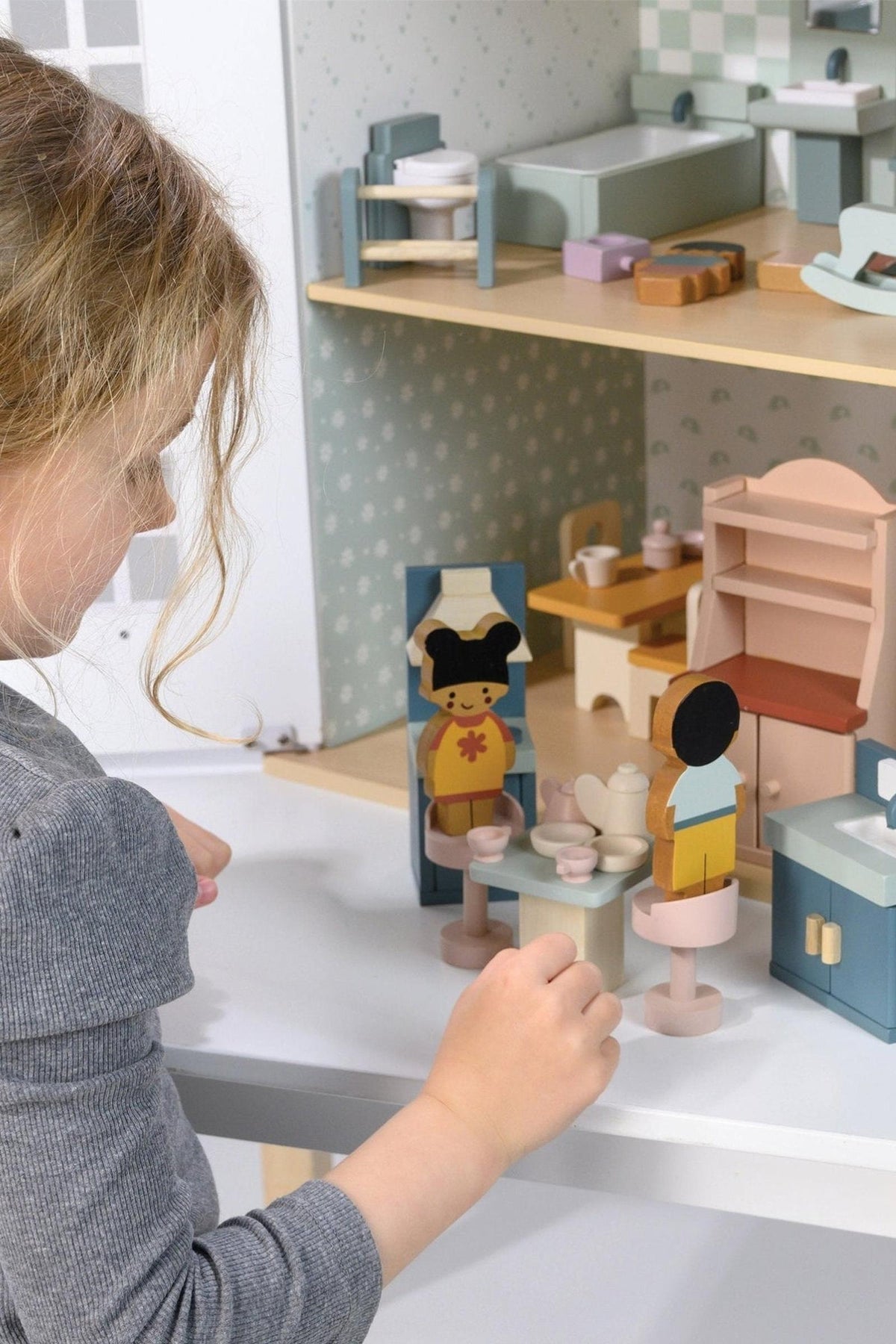 A child interacts with colorful wooden dolls and furniture in a dollhouse setting. The scene features a kitchen area with a table, chairs, and a mirror, highlighting imaginative play.