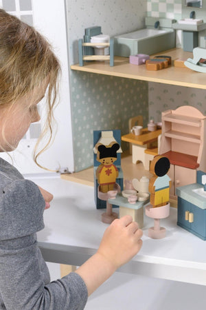 A child interacts with colorful wooden dolls and furniture in a dollhouse setting. The scene features a kitchen area with a table, chairs, and a mirror, highlighting imaginative play.