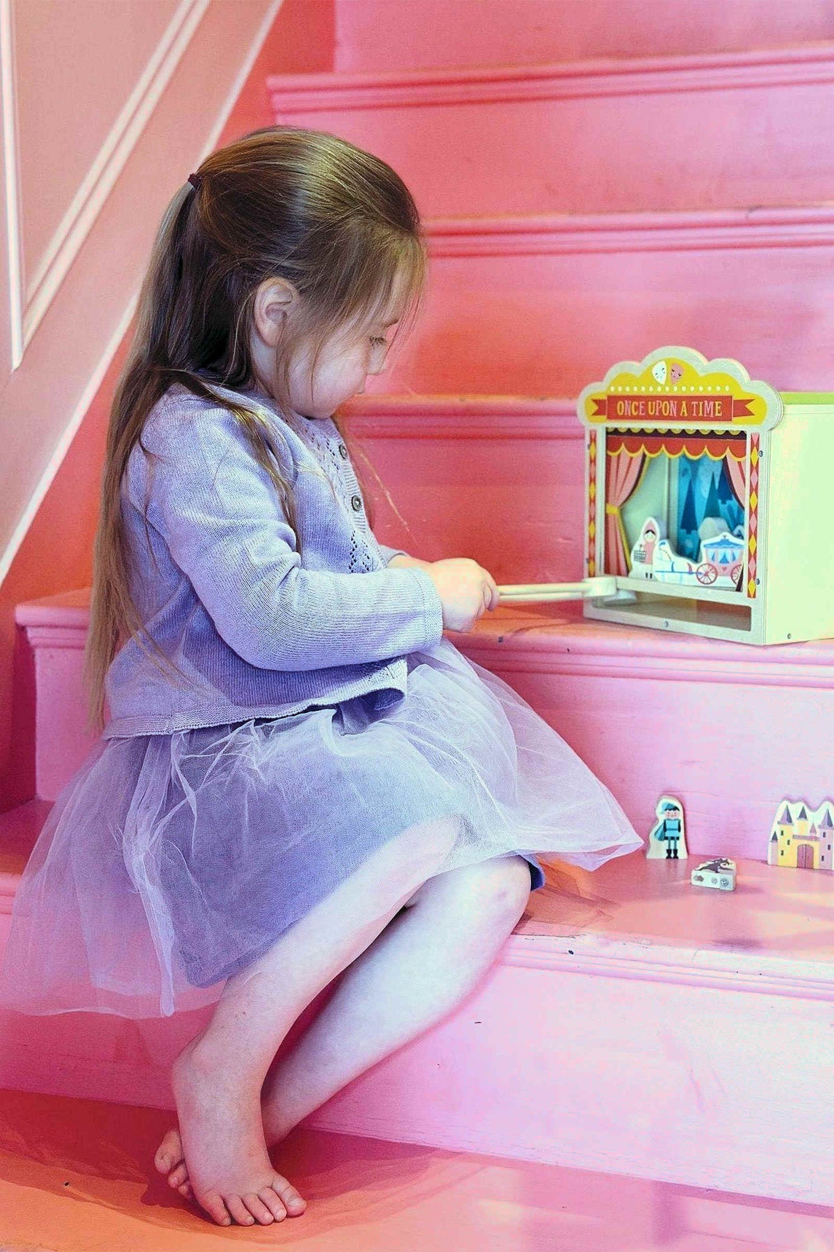A child in a purple dress plays with a wooden tabletop theatre on pink stairs, featuring small toy figures.