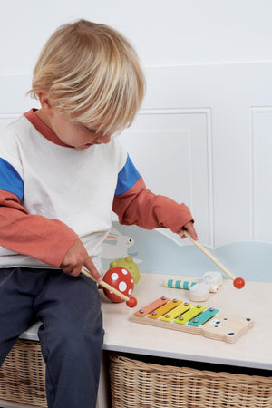 A young child plays with a wooden bear xylophone and colorful percussion instruments on a light wooden table.