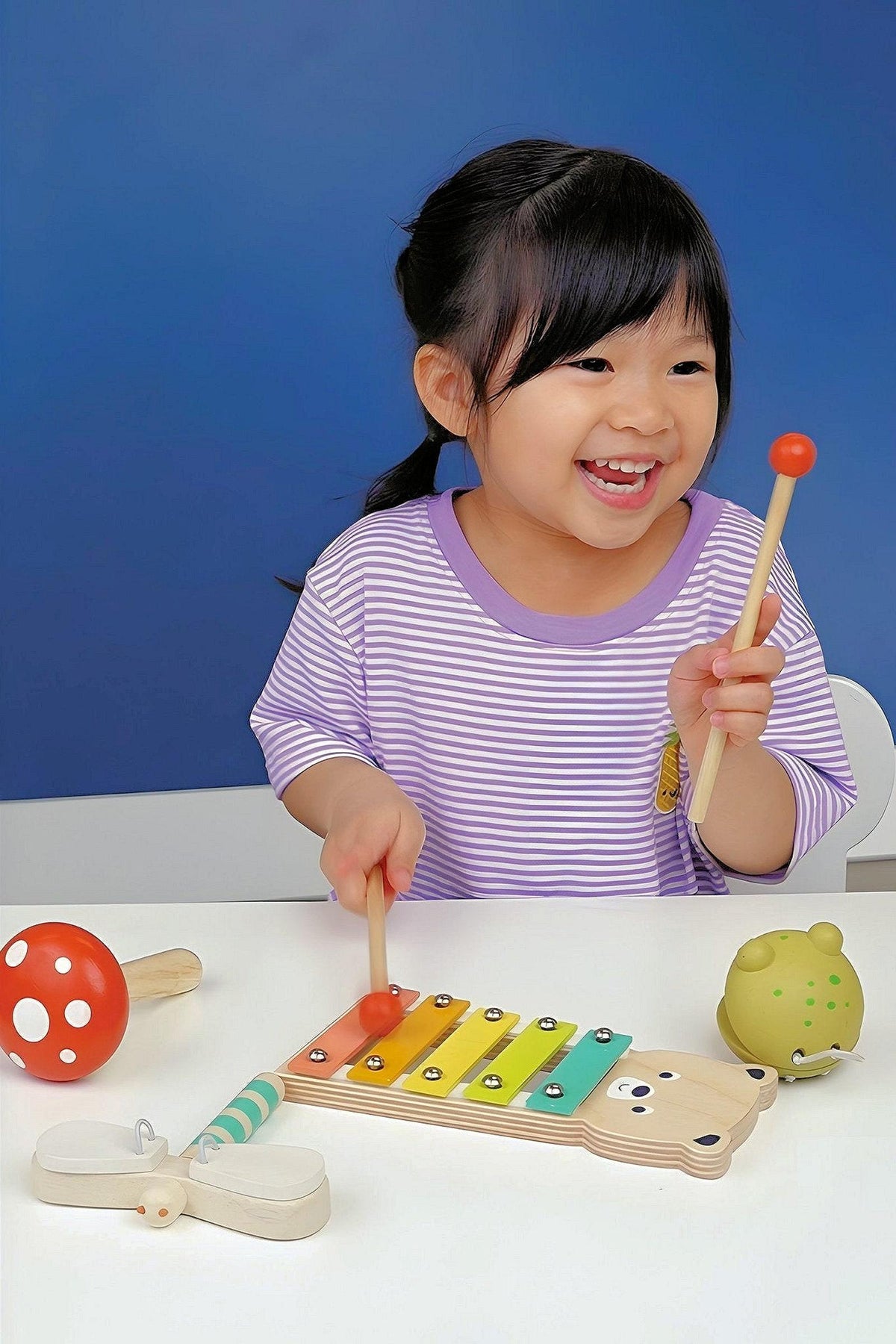 A cheerful child plays with a wooden bear xylophone, using mallets to strike colorful keys. Nearby, a dragonfly clacker and a toadstool maraca add to the whimsical musical scene.