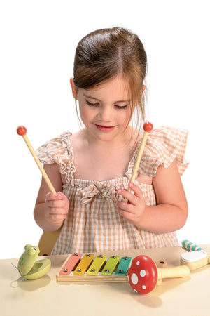 A young girl in a checked dress plays with a wooden xylophone, holding mallets. The colorful xylophone features a toadstool maraca and froggy castanet nearby, promoting musical exploration.