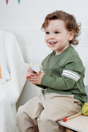 A smiling child with curly hair sits on a wooden bench, holding a dragonfly clacker. He wears a green long-sleeve shirt with white stripes, exuding joy in a cozy room.
