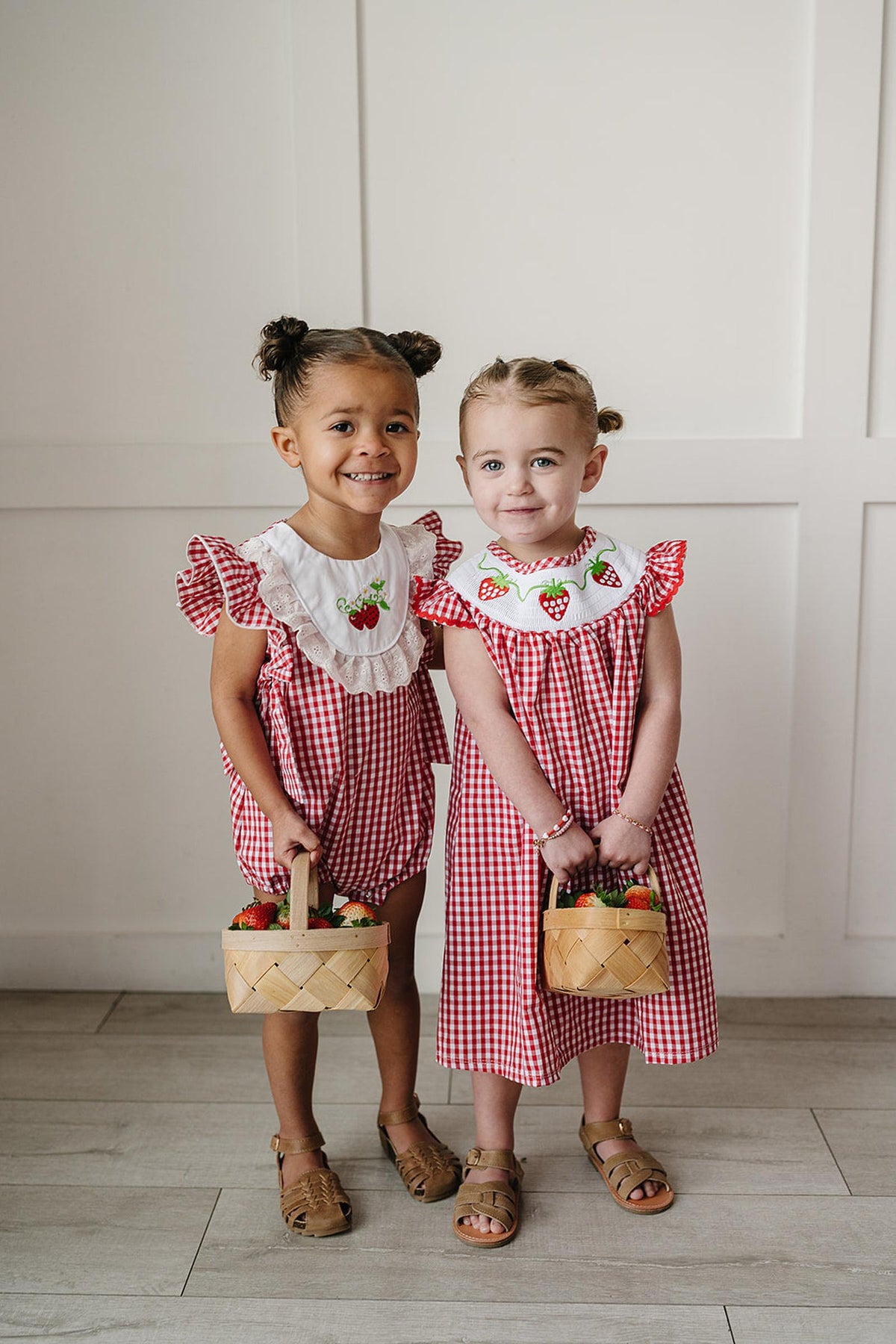 Two young girls are smiling while wearing adorable red gingham dresses with embroidered strawberries on the neckline.