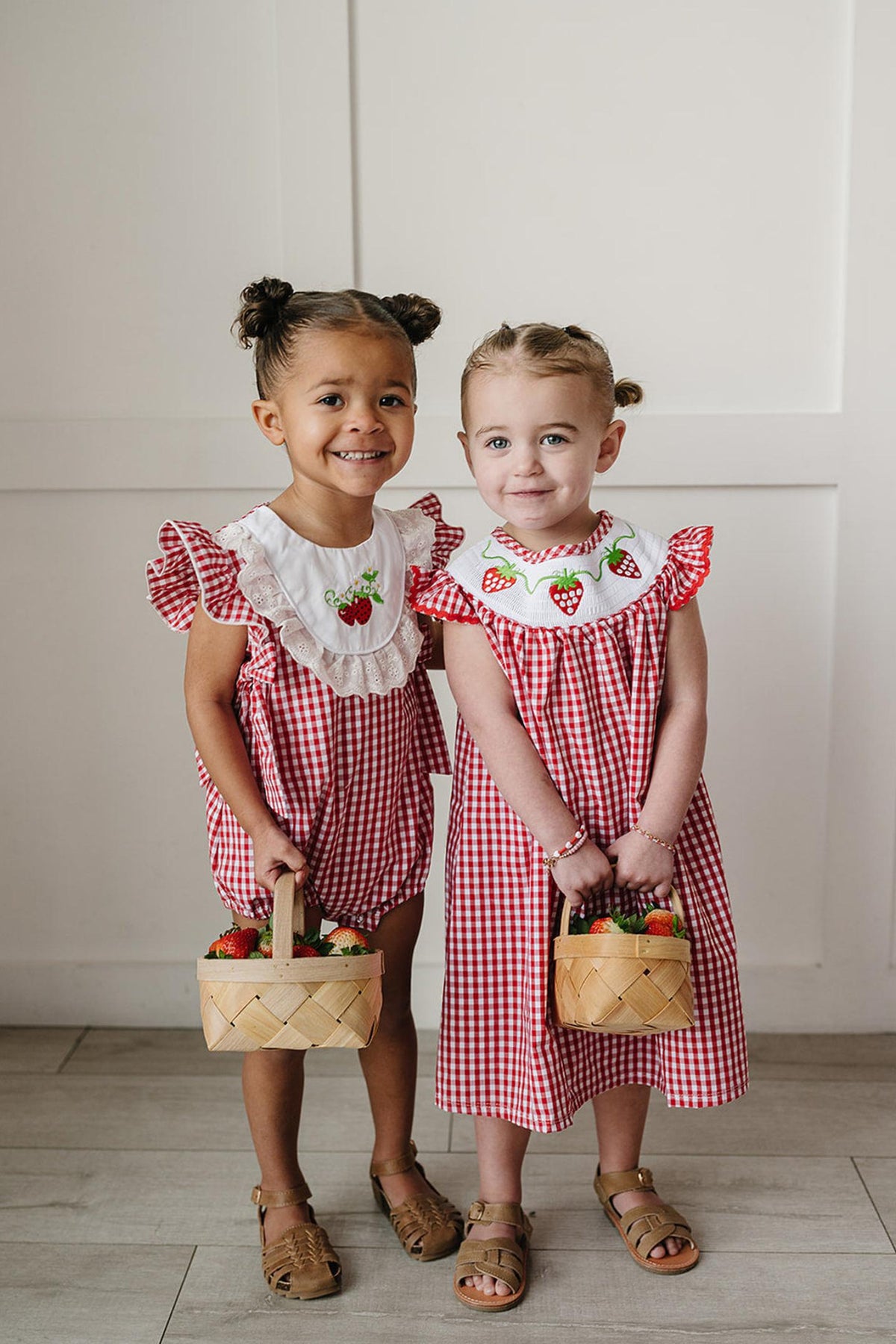Two young girls stand side by side, smiling while wearing red and white gingham outfits with ruffle sleeves.