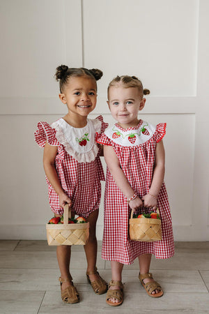 Two young girls stand side by side, smiling while wearing red and white gingham outfits with ruffle sleeves.