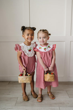 Two young girls are smiling while wearing adorable red gingham dresses with embroidered strawberries on the neckline.