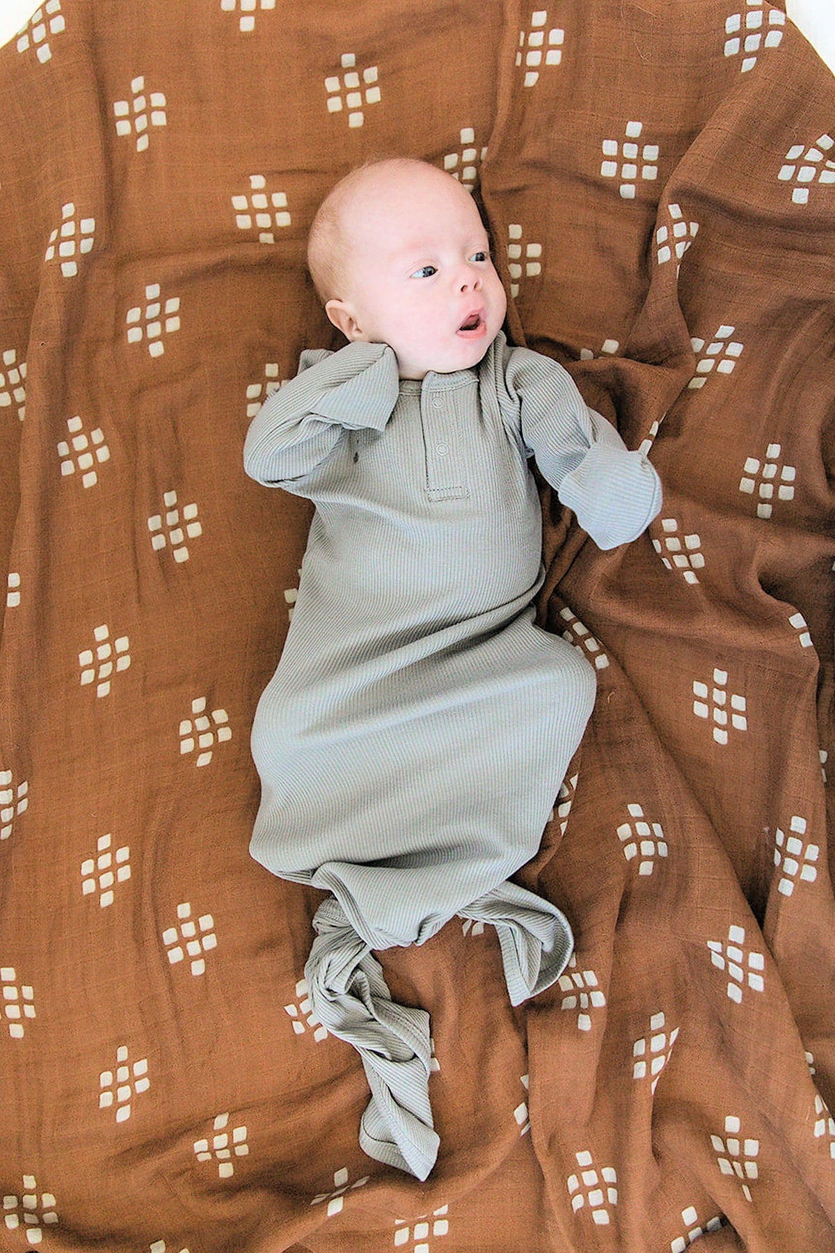A baby wearing a grey ribbed knot gown lies on a brown patterned blanket, looking up.