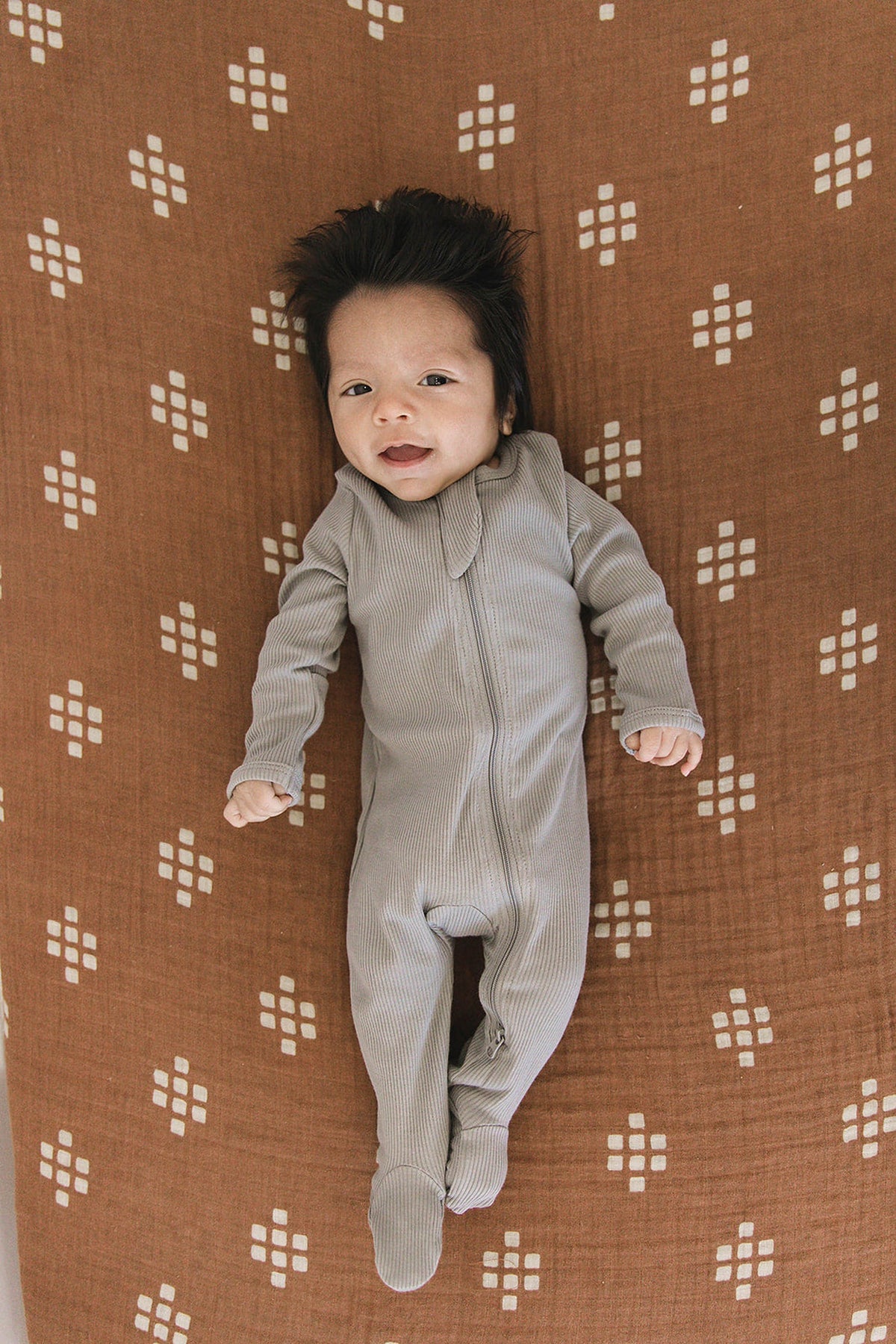 A baby in a grey ribbed footed pajama lies on a brown patterned blanket, smiling contently.