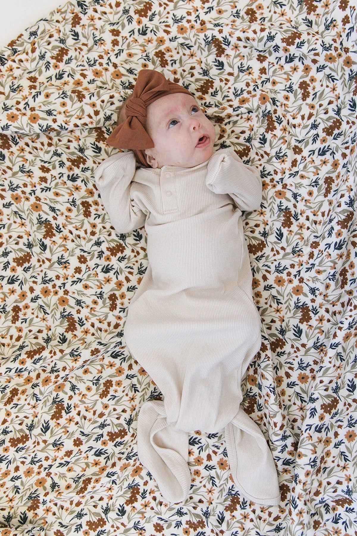 A baby in a vanilla ribbed gown with a brown bow lies on a floral-patterned blanket.