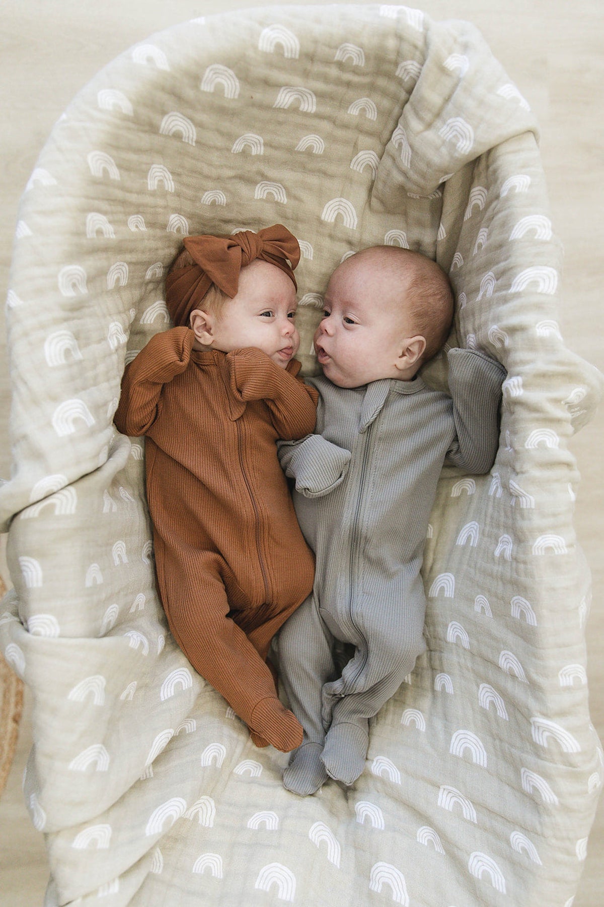 Two babies in ribbed zipper footed pajamas, one in brown, the other in grey, lying on a patterned blanket.